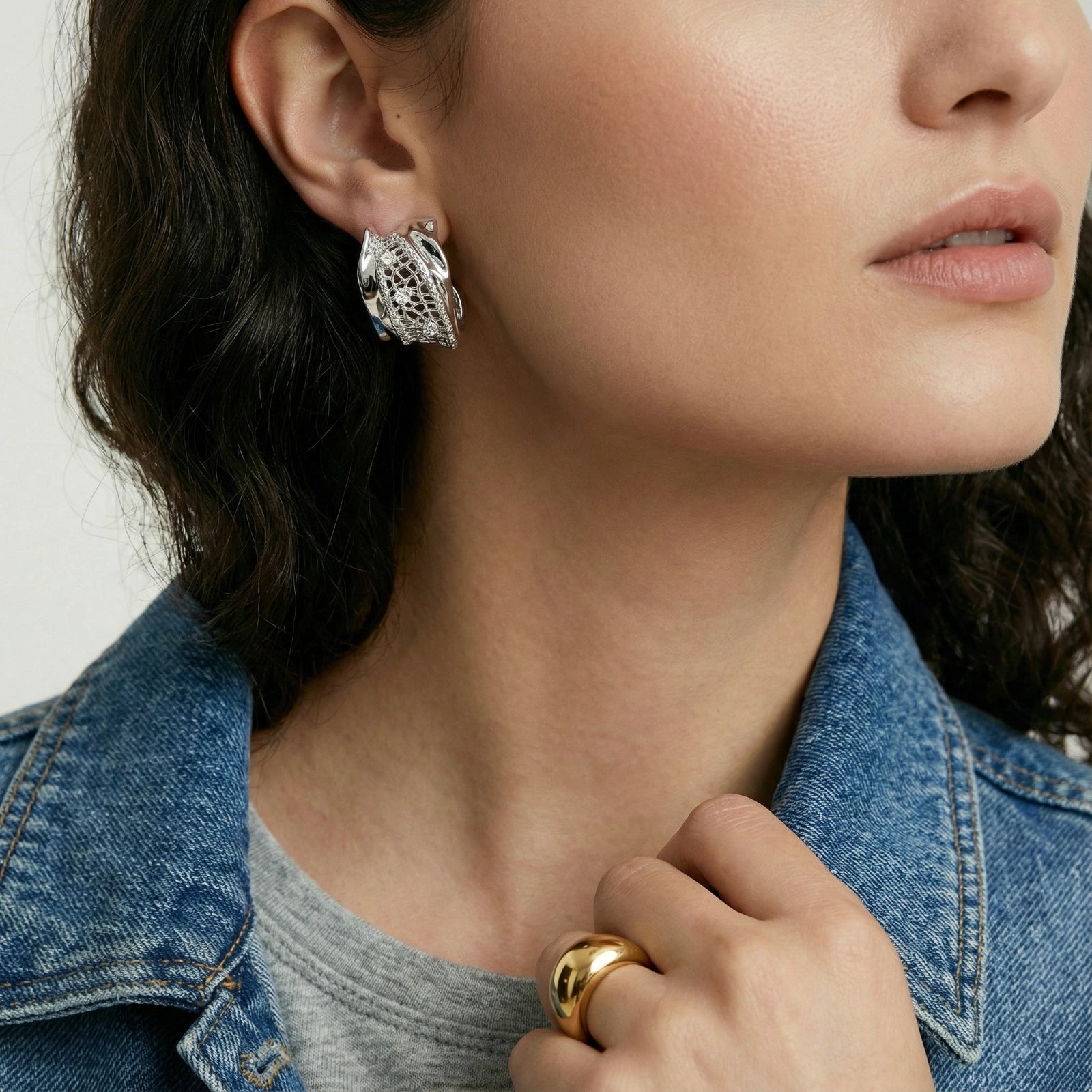 Close-up of a woman wearing silver earrings and a gold ring, with a denim jacket.