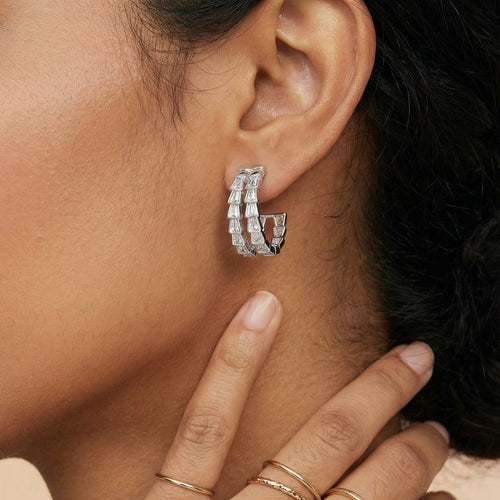 Close-up of a person wearing silver hoop earrings and gold rings on a neutral background