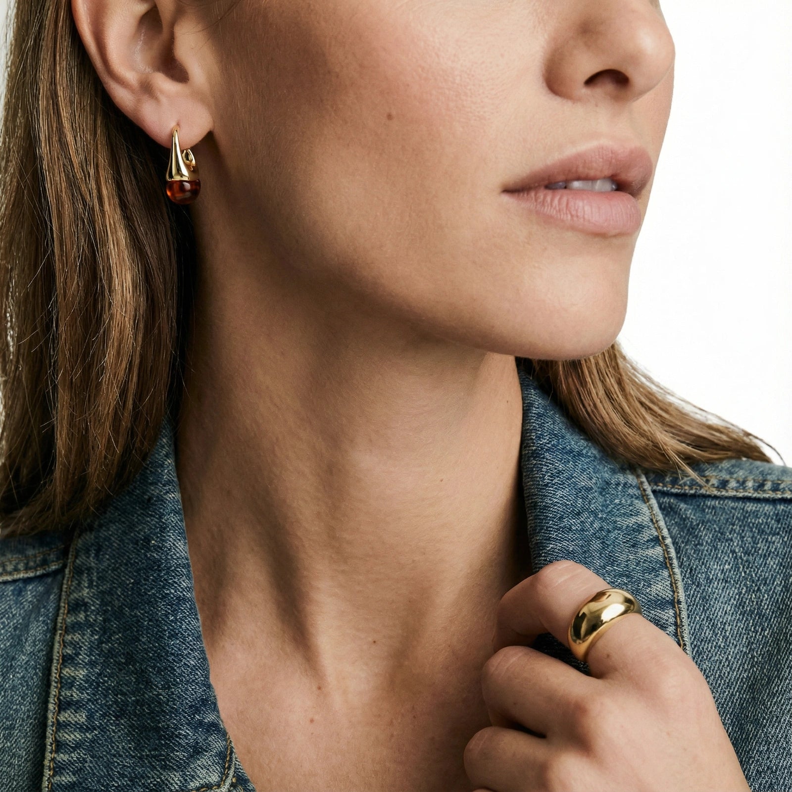 Close-up of a woman wearing gold earrings and a ring, with a denim jacket.