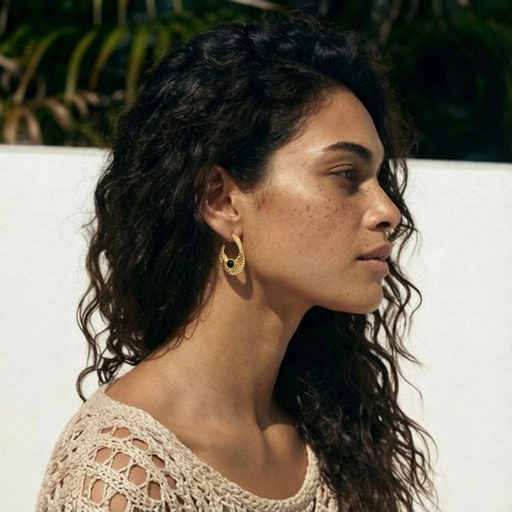 Woman with long dark hair wearing gold hoop earrings, sitting outdoors.