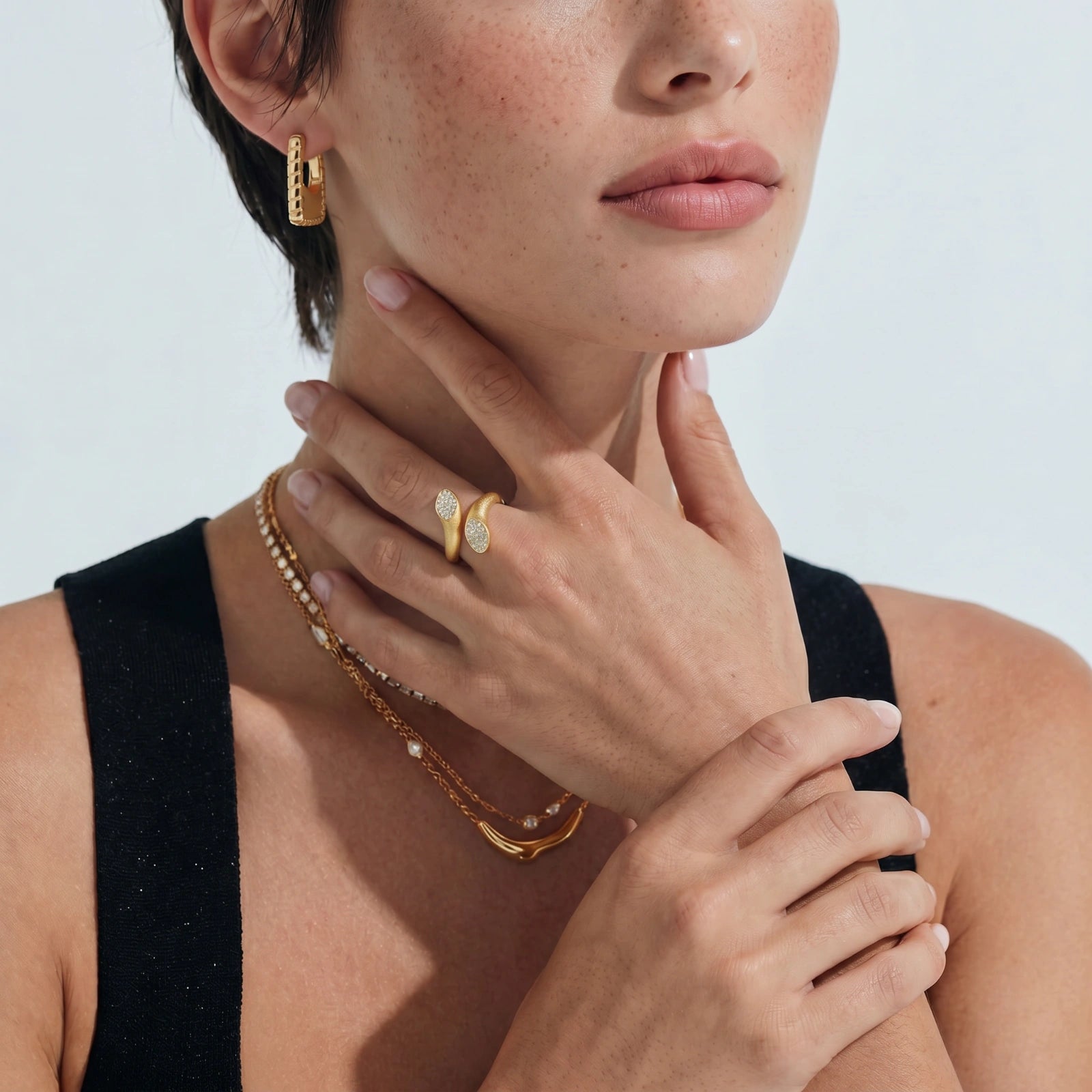 Close-up of a woman wearing gold jewelry including earrings, necklace, and ring on a light background