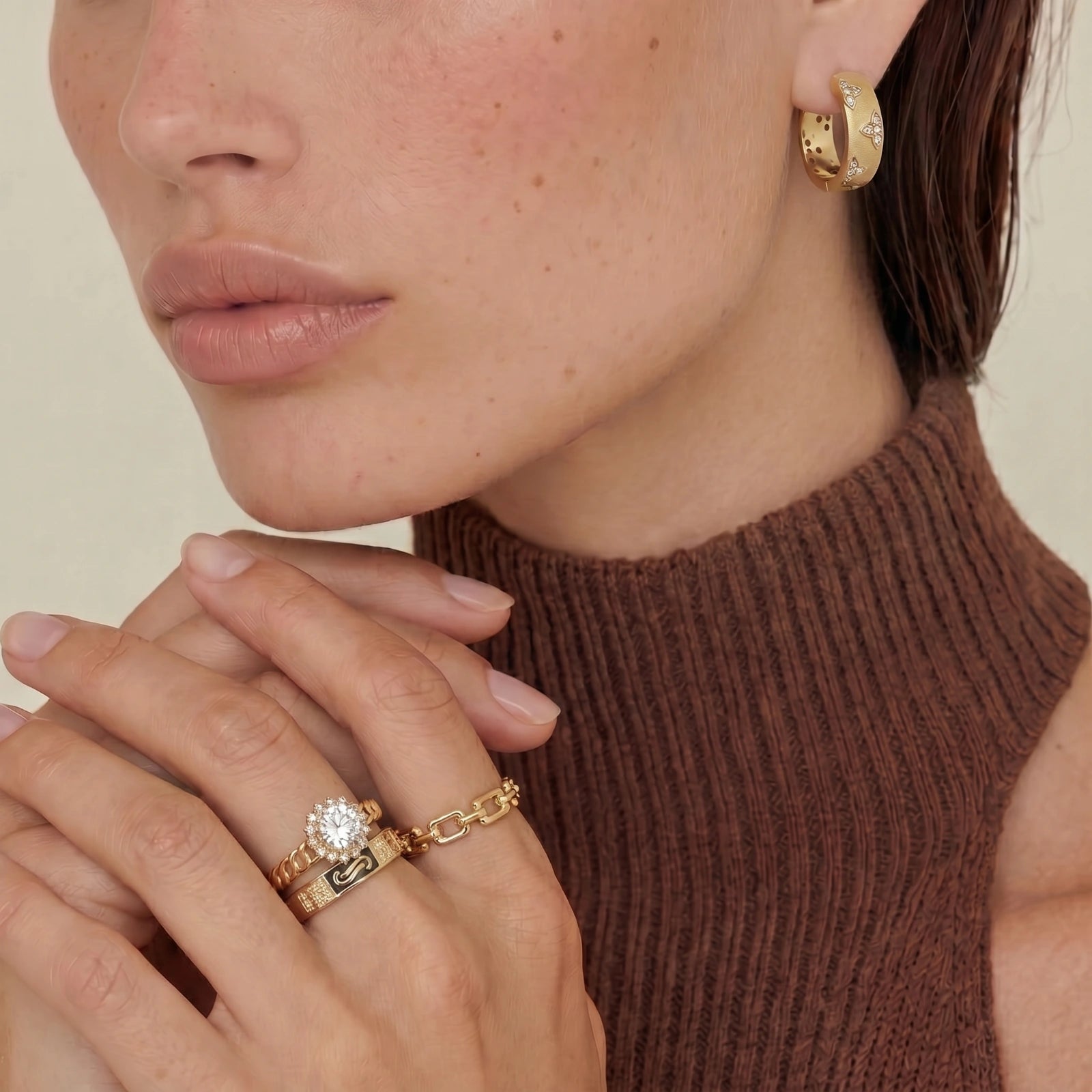 Close-up of a person wearing gold hoop earrings and rings on a neutral background